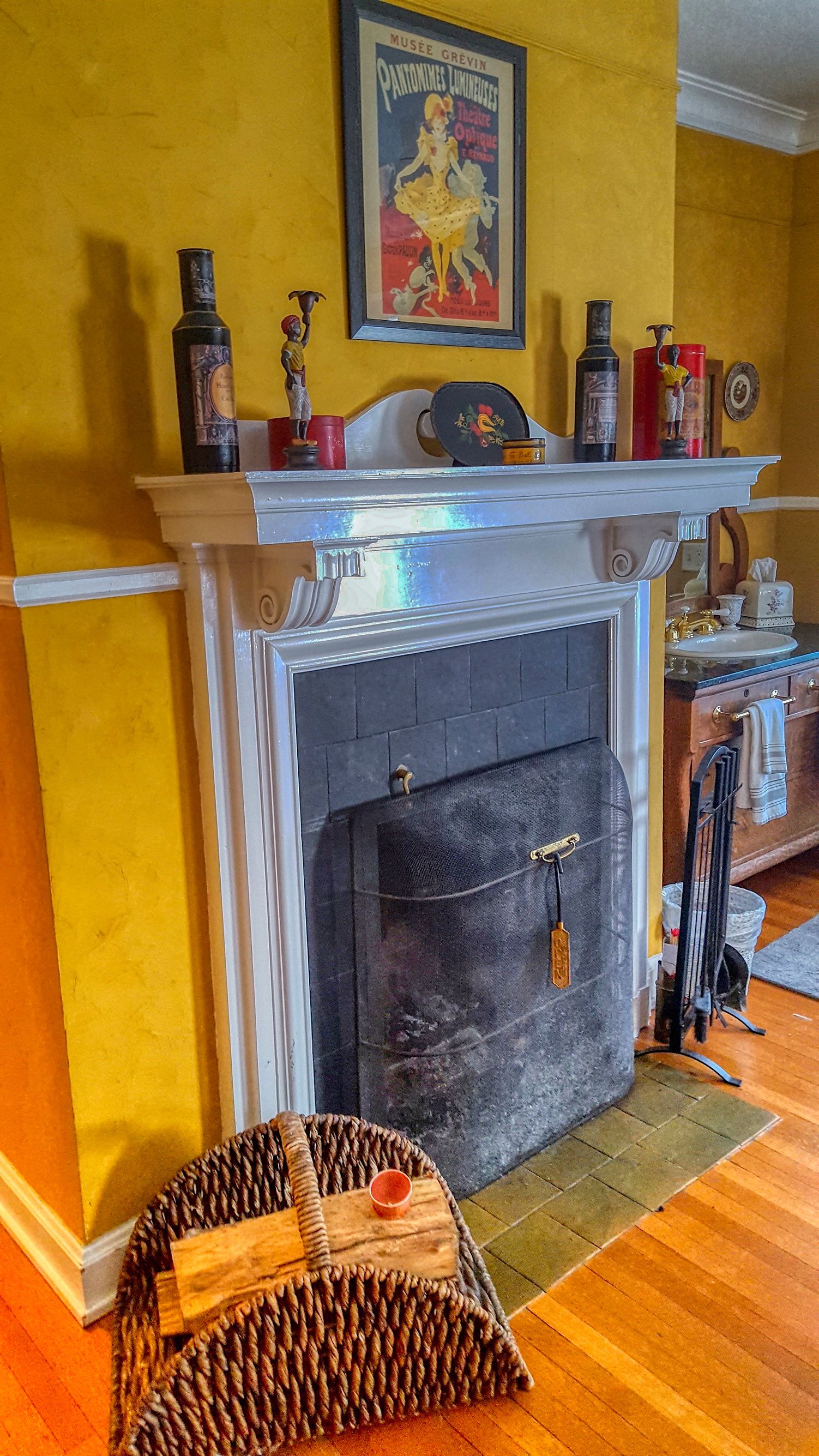Una habitación con chimenea, un repisa decorada con adornos, un cartel enmarcados en la pared y una cesta tejida en el suelo.