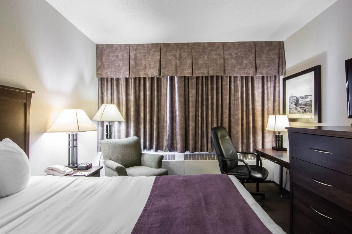 Habitación de hotel con cama, dos lámparas de noche, un escritorio con silla, cómoda y un amplio ventanal con cortinas.