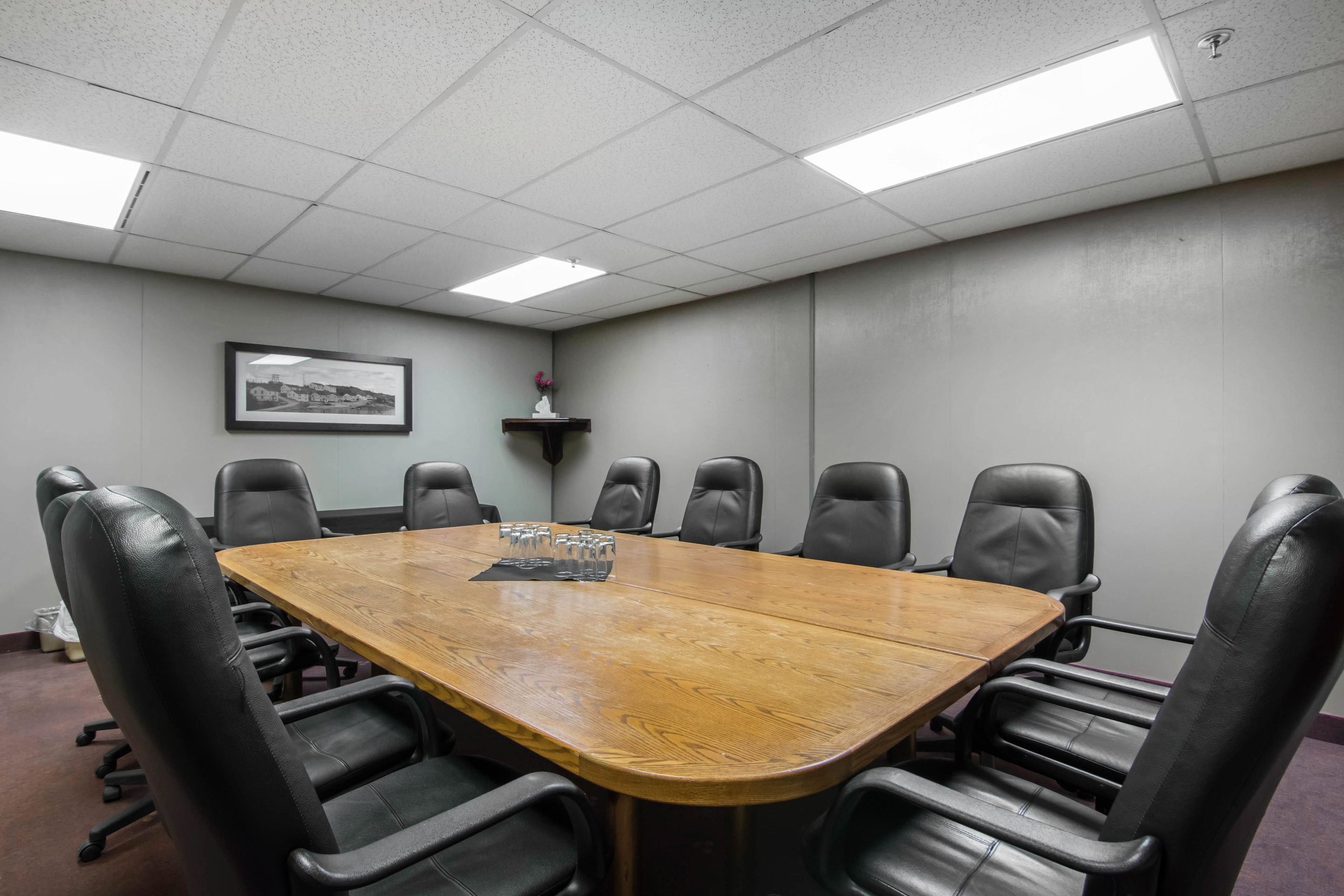 A conference room with a large wooden table surrounded by black leather chairs, a framed picture on the wall, and a small shelf with a vase.