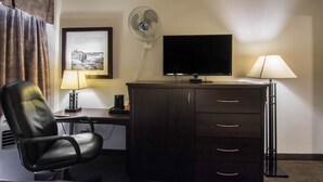 A hotel room with a desk, a chair, a television, a lamp, a fan, and a cabinet.