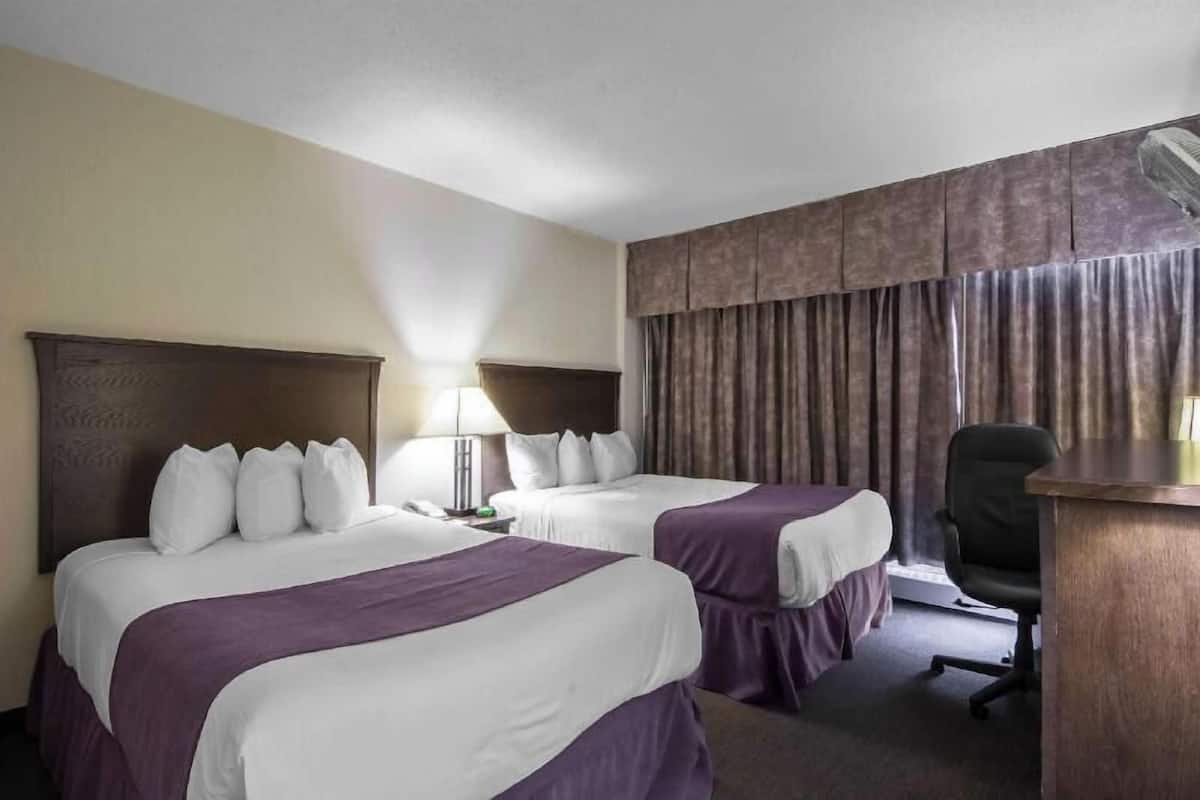 Habitación de hotel con dos camas, un escritorio, una silla, una lámpara y una ventana con cortinas.