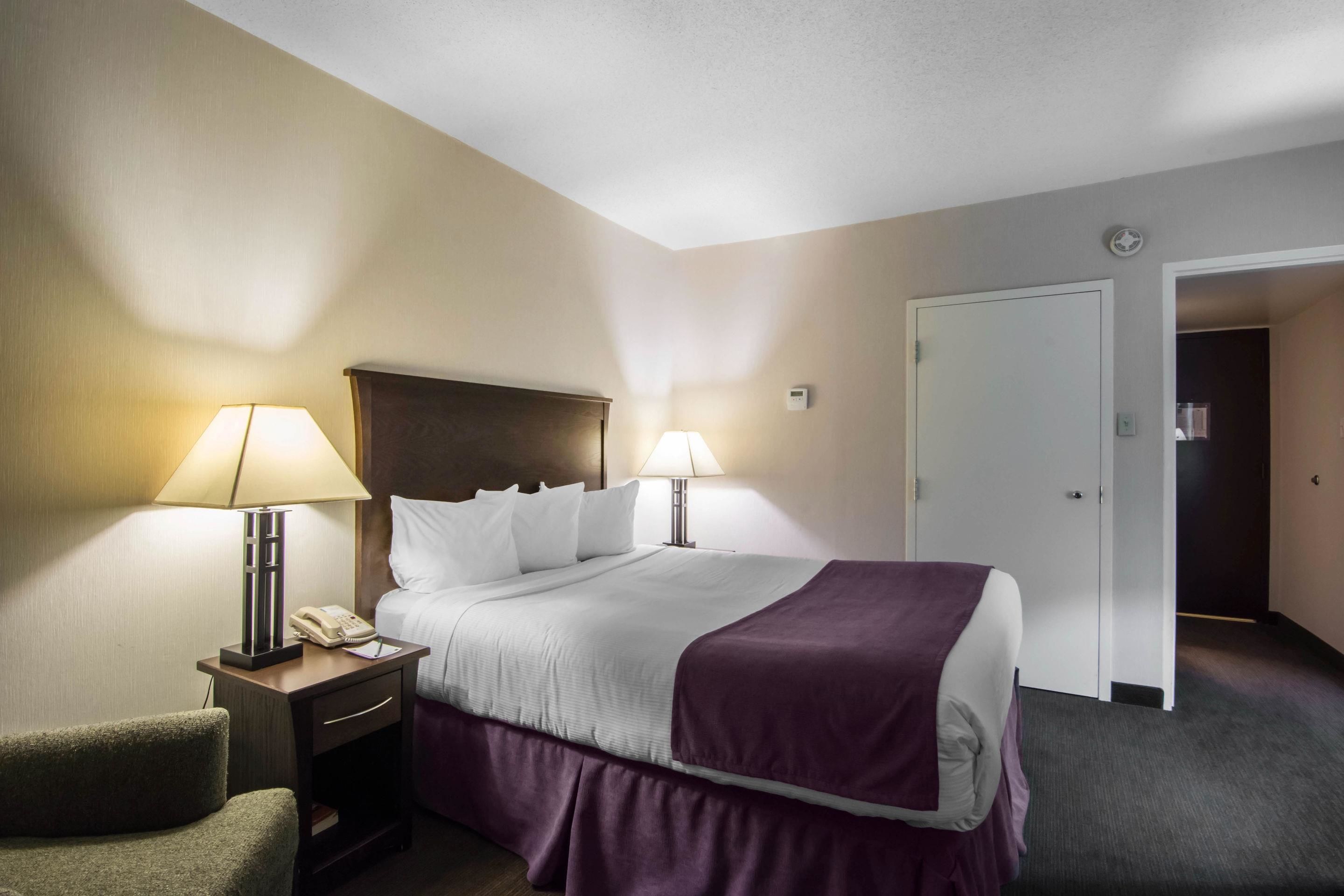 A hotel room with a bed, two bedside lamps, a nightstand with a phone, a chair, and a door leading to another room.