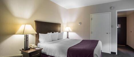 A hotel room with a bed, two bedside lamps, a nightstand with a phone, a chair, and a door leading to another room.