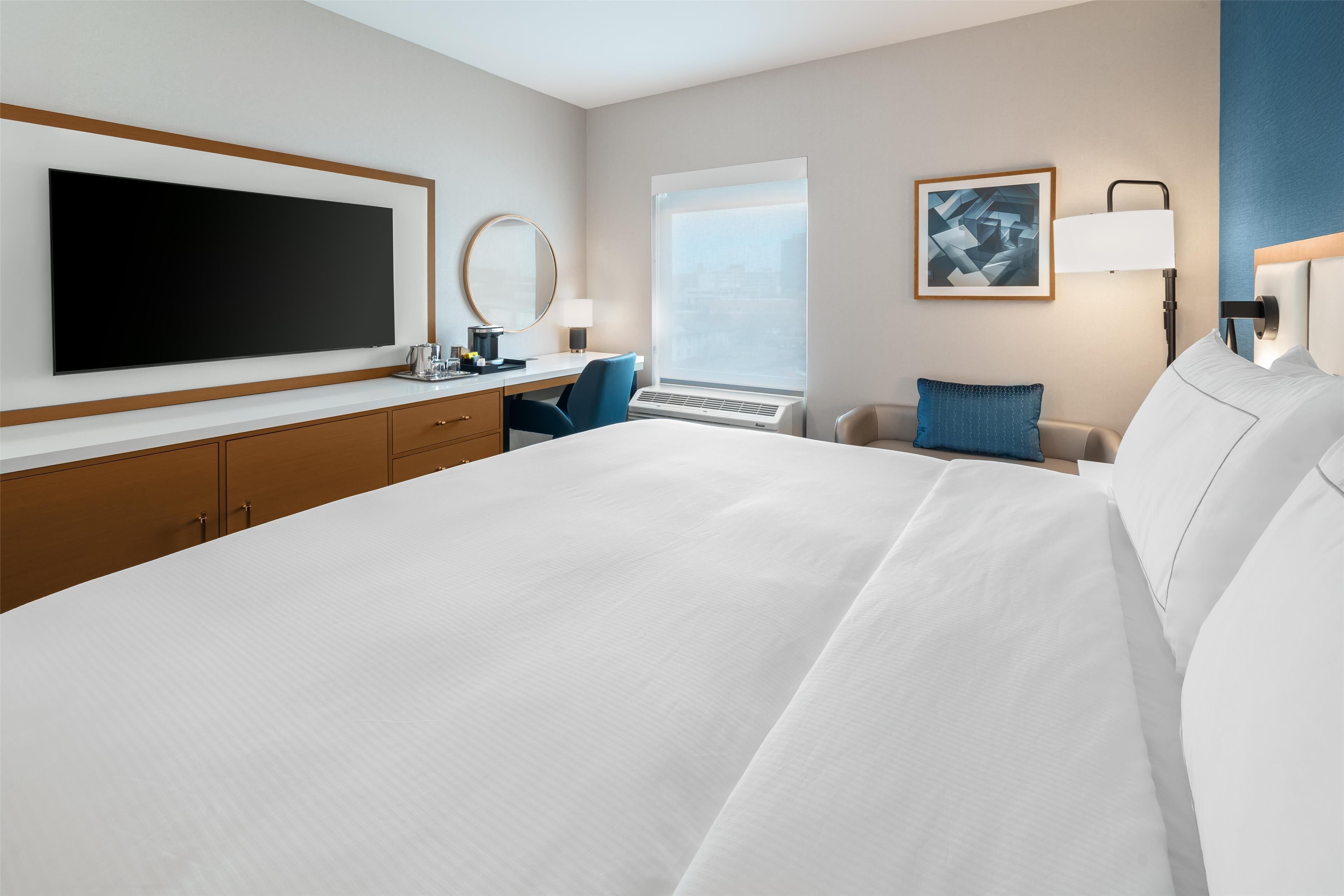 Quarto de hotel com uma cama grande, uma mesa com cadeira, televisão de tela plana, uma janela ampla e um quadro na parede.
