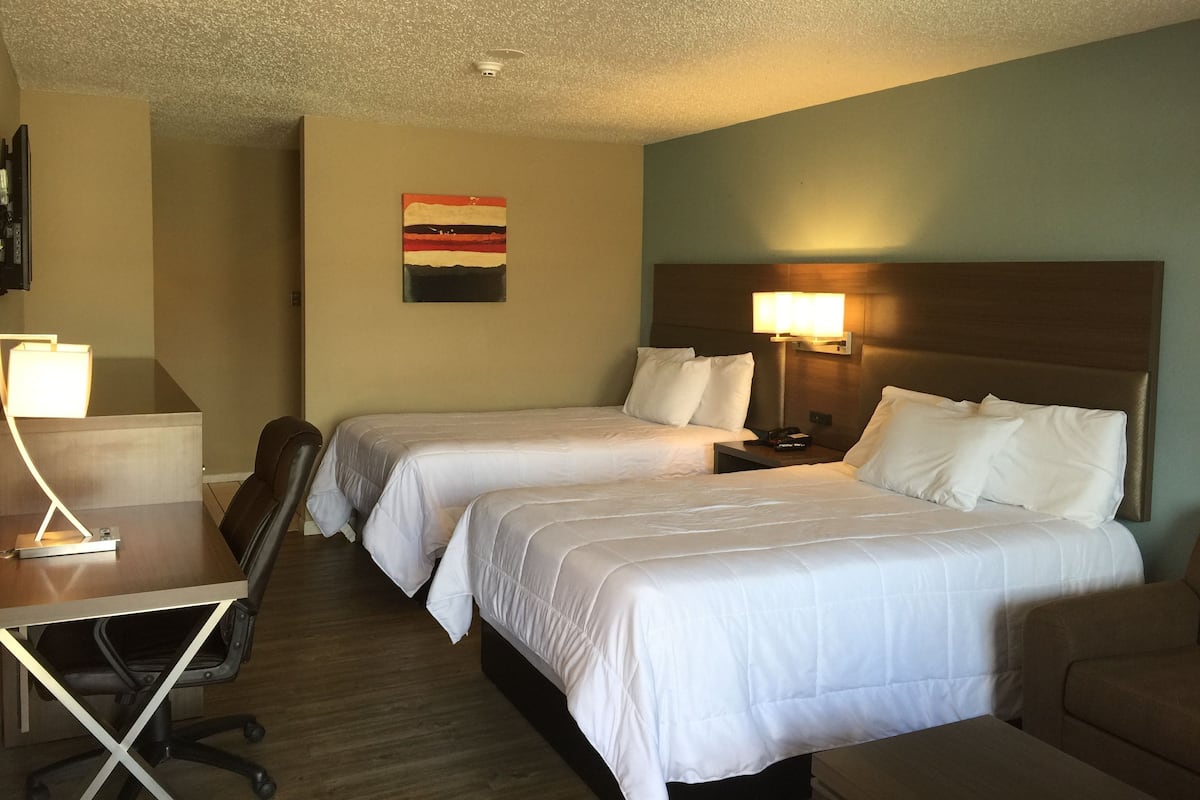 A hotel room with two beds, a desk, a chair, a lamp, and a painting on the wall.