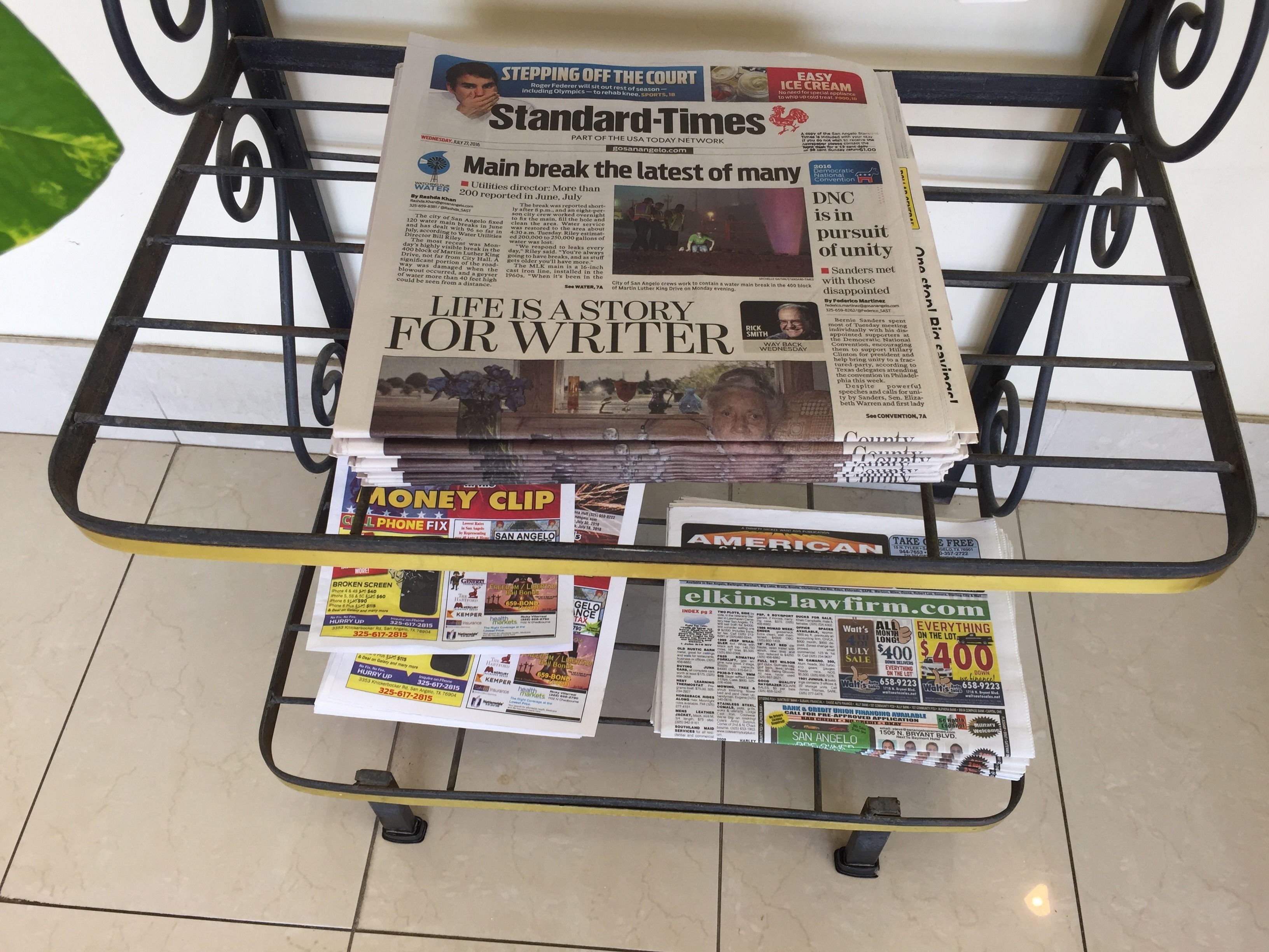 A newspaper rack with various newspapers, including the Standard Times, and a few flyers on a glass table.
