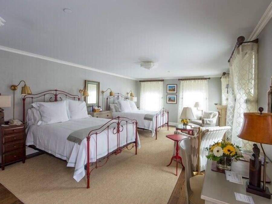Quarto de hotel com duas camas, uma escrivaninha, uma cadeira e uma mesinha de cabeceira com abajur e flores.