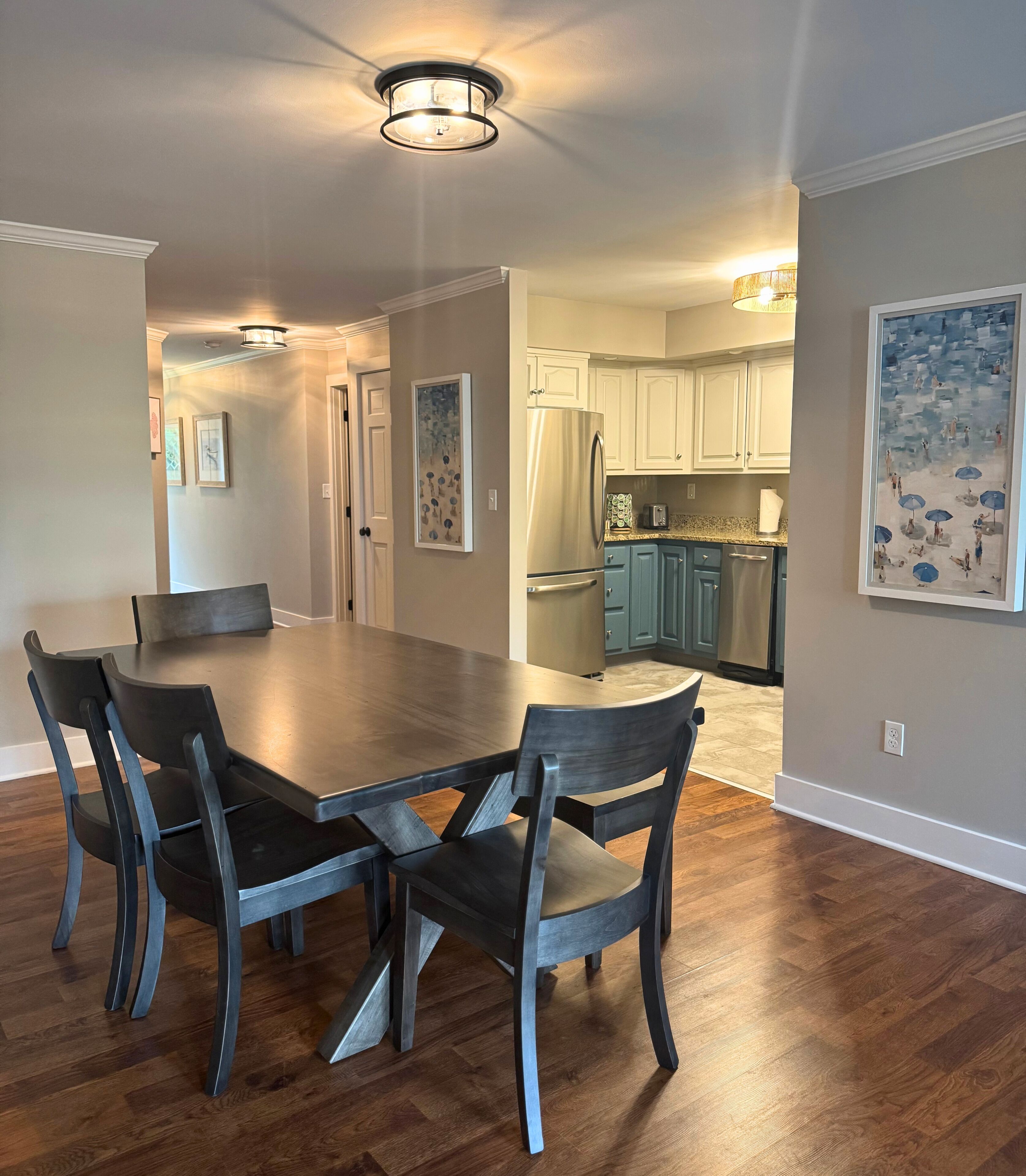 A dining area with a large table and chairs, a kitchen in the background, and a ceiling light fixture.