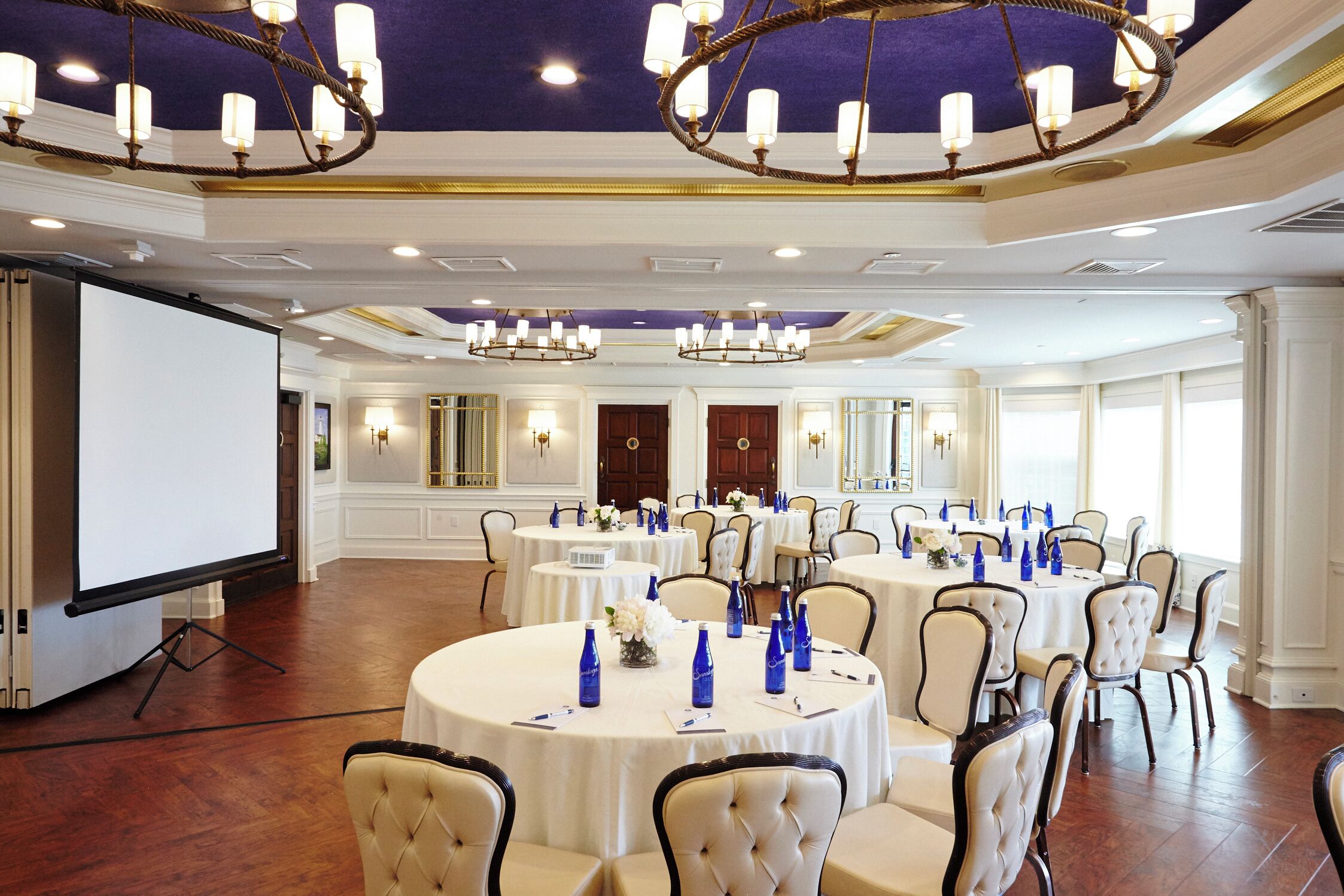 A spacious banquet hall with multiple round tables, each set with white tablecloths and blue water bottles.
