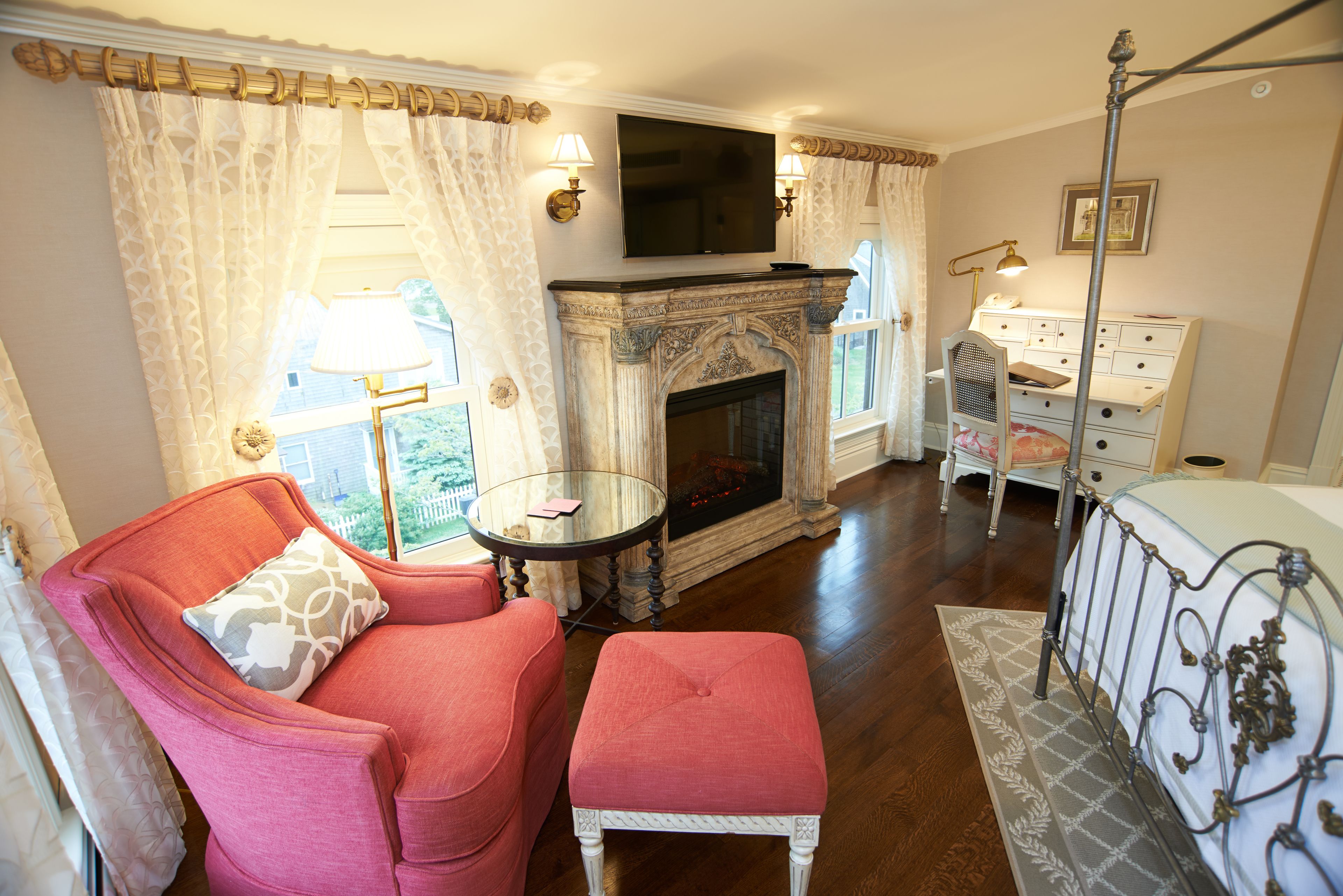 A room with a fireplace, a television mounted above it, a pink armchair, a small table, and a white dresser.
