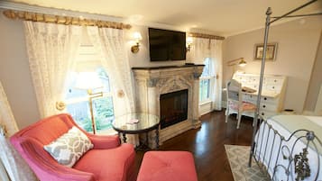 A room with a fireplace, a television mounted above it, a pink armchair, a small table, and a white dresser.
