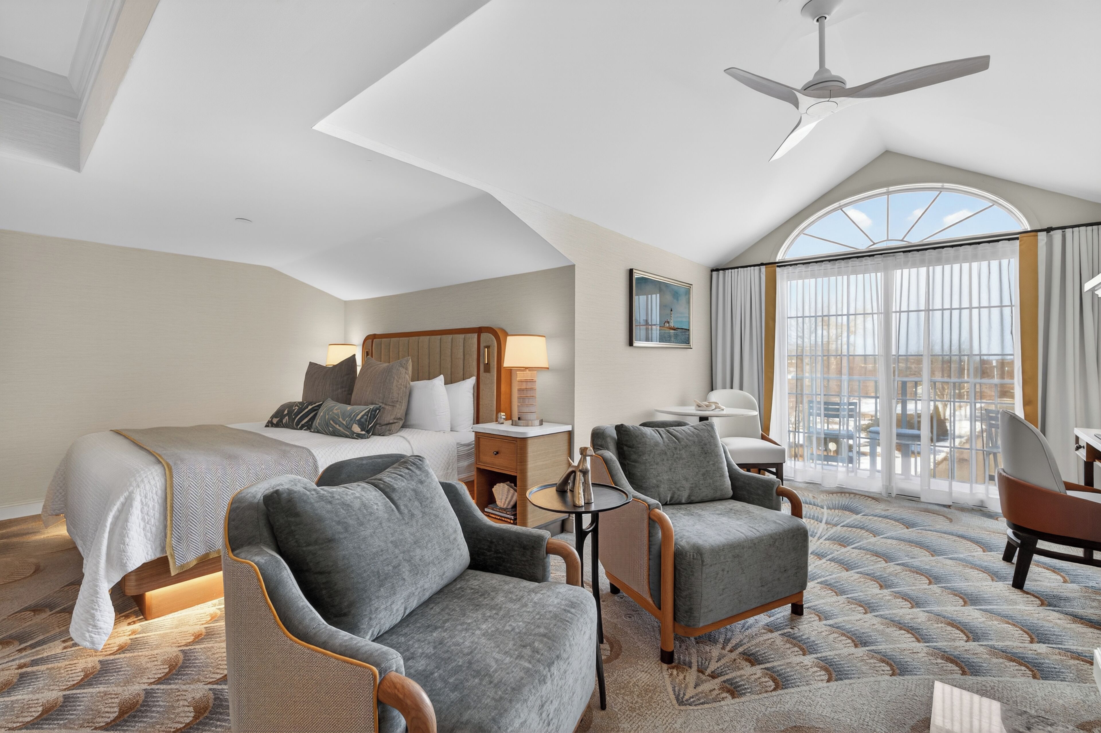 A hotel room with a bed, two armchairs, a small table, a ceiling fan, and a large window with curtains.