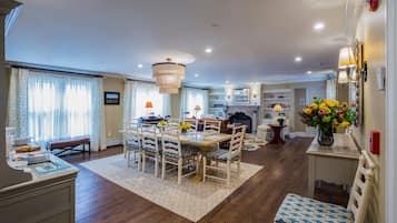 A dining area with a wooden table, chairs, and a chandelier. There is a fireplace, a rug, and a vase with flowers.