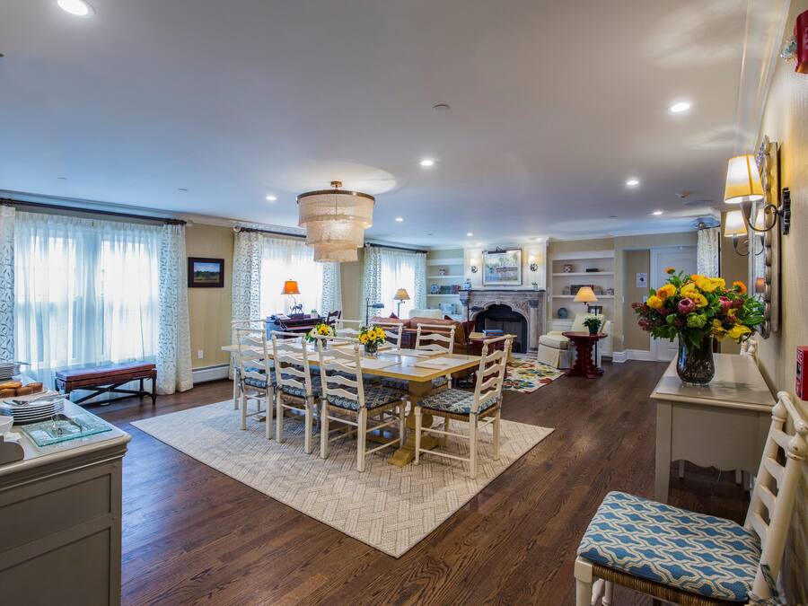 A dining area with a wooden table, chairs, and a chandelier. There is a fireplace, a rug, and a vase with flowers.