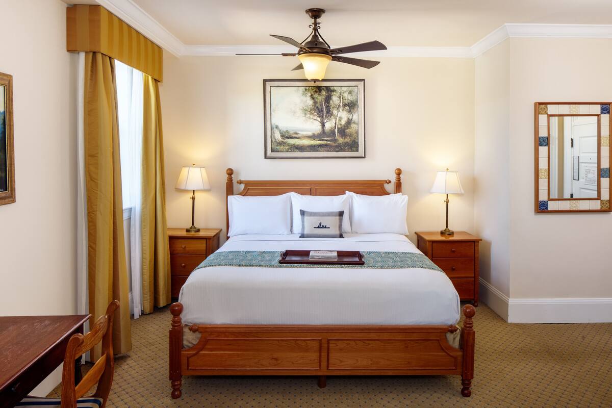 A neatly arranged hotel room with a wooden bed, two bedside tables with lamps, a ceiling fan, a framed landscape painting, and a large window with curtains.