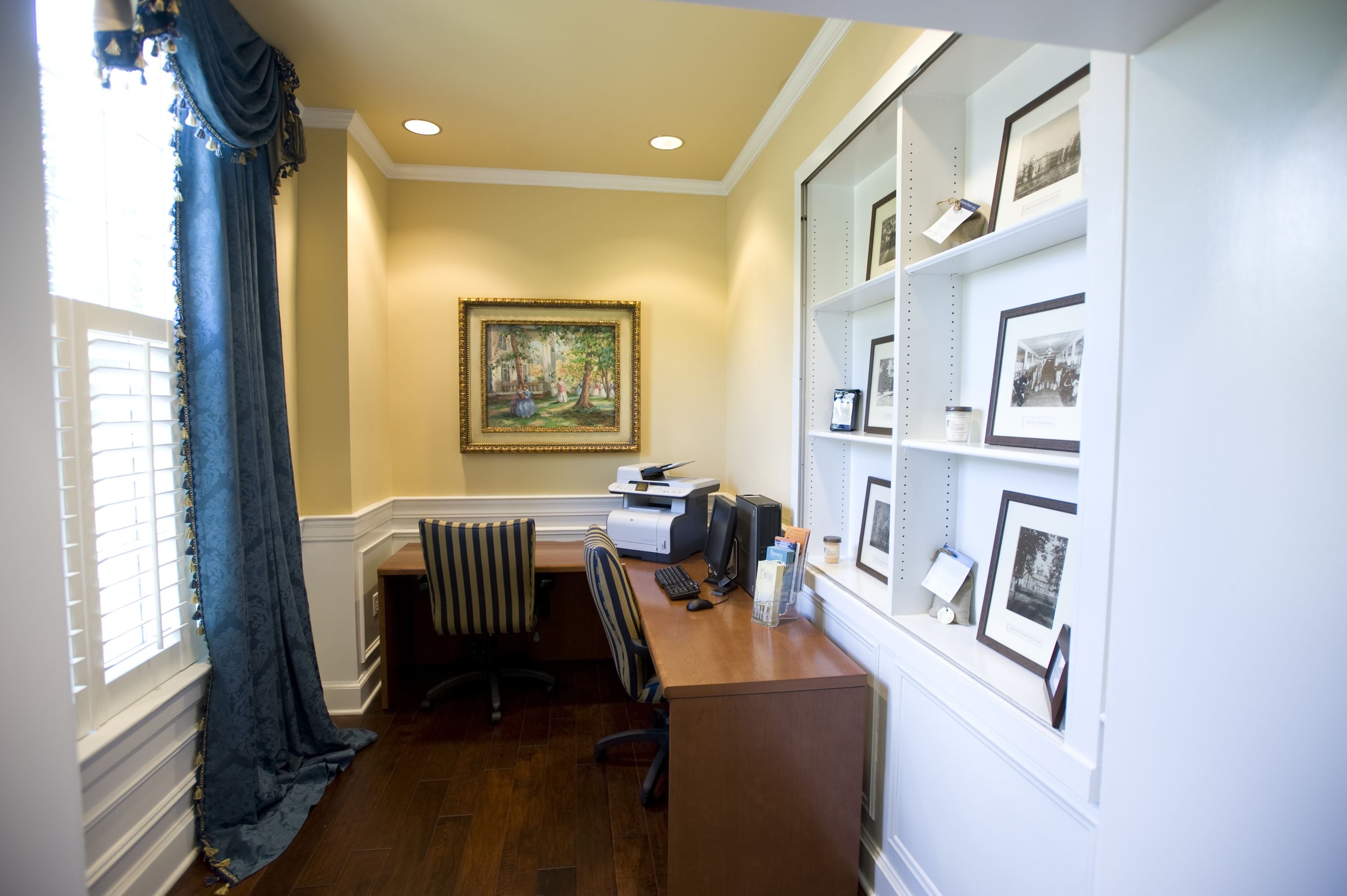 A home office with a desk, computer, printer, and framed pictures on shelves.
