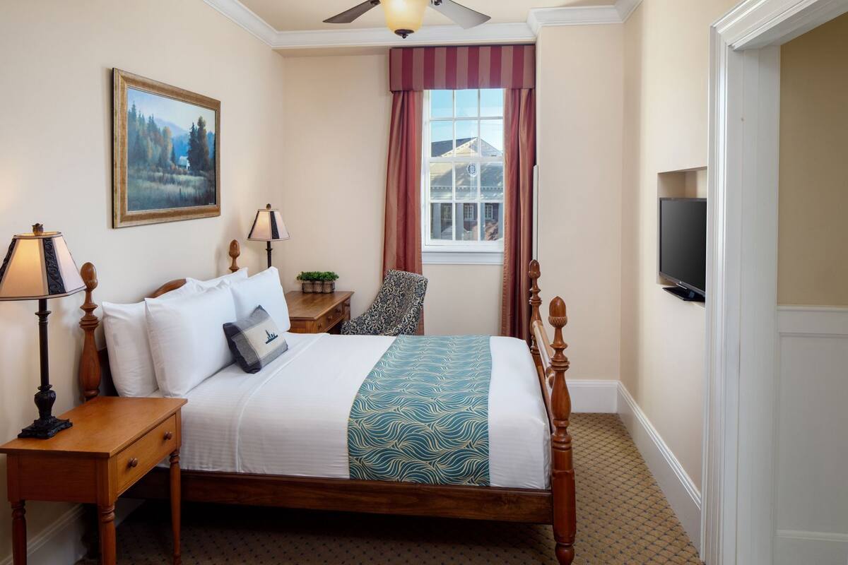A bedroom with a bed, bedside tables, a lamp, a chair, a television, a painting, and a window with curtains.