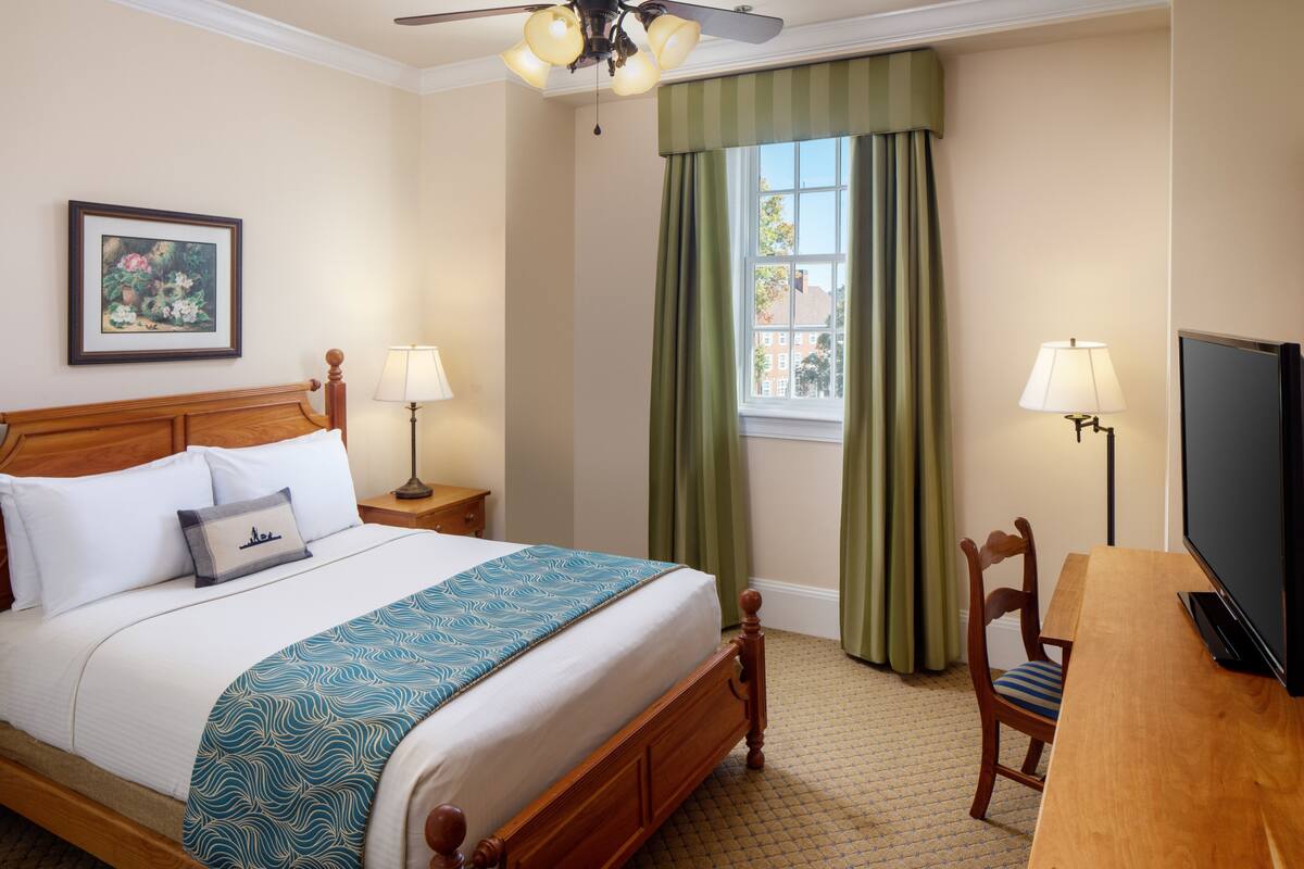 A hotel room with a bed, two bedside lamps, a television, a window with curtains, and a framed picture on the wall.