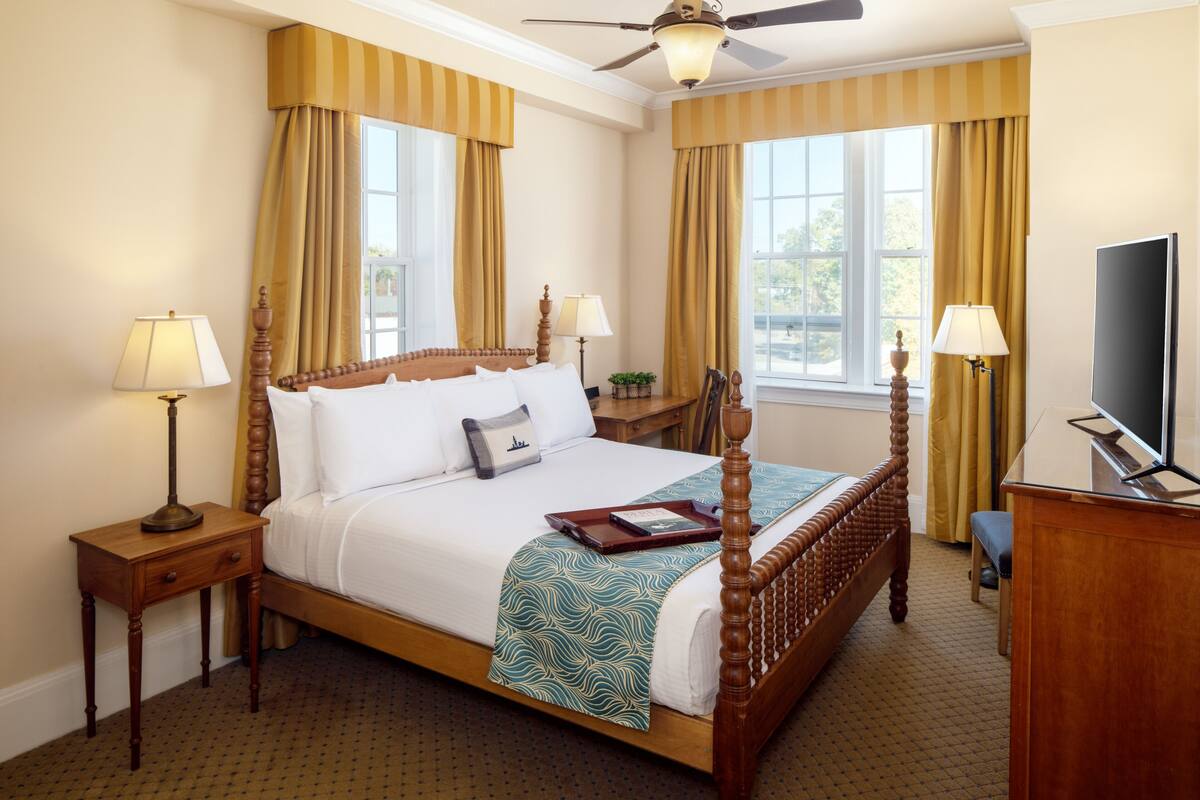 A hotel room with a four-poster bed, two bedside tables with lamps, a desk with a chair, and a television.