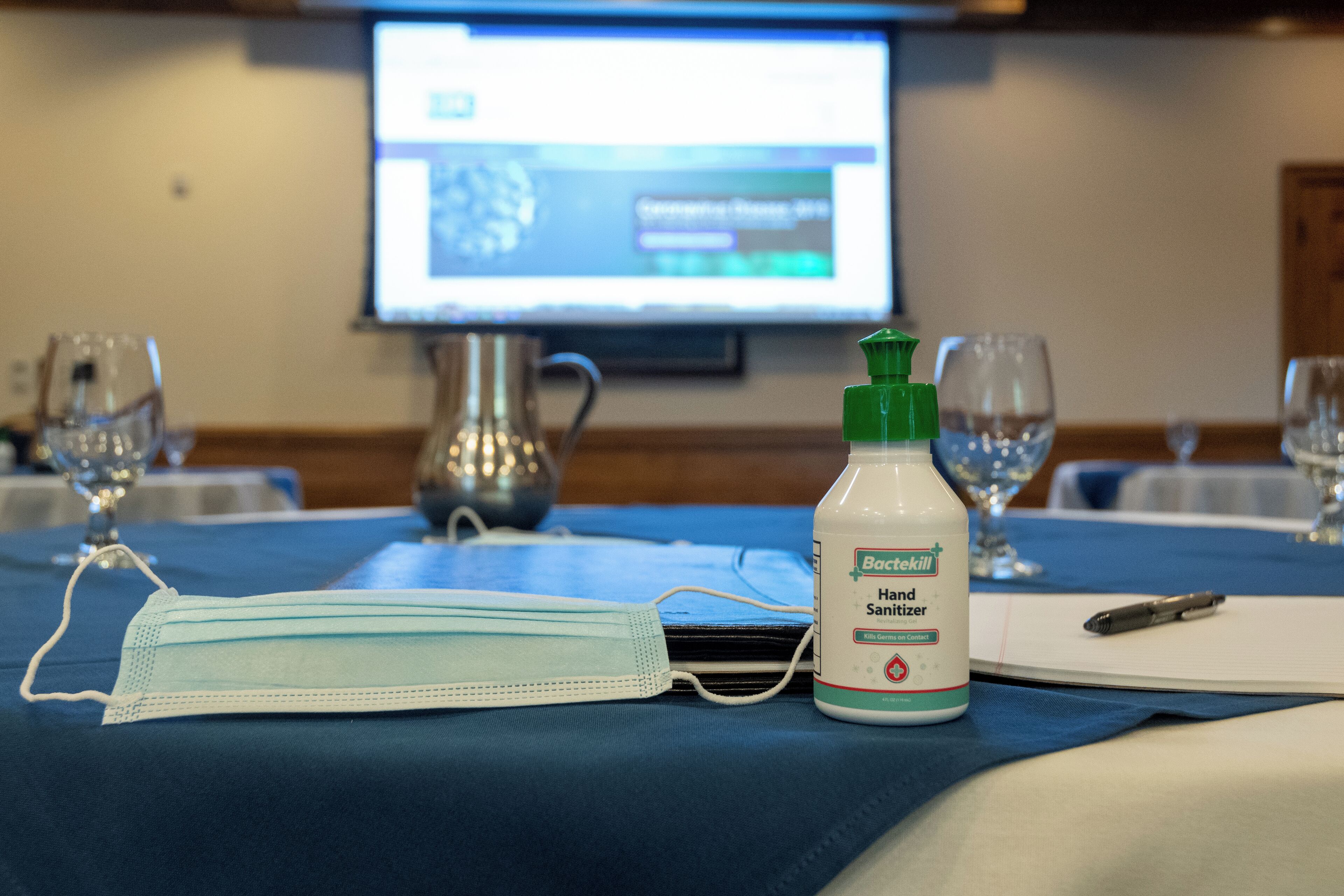 A hotel room with a table set for a meeting, a face mask, and a bottle of hand sanitizer.