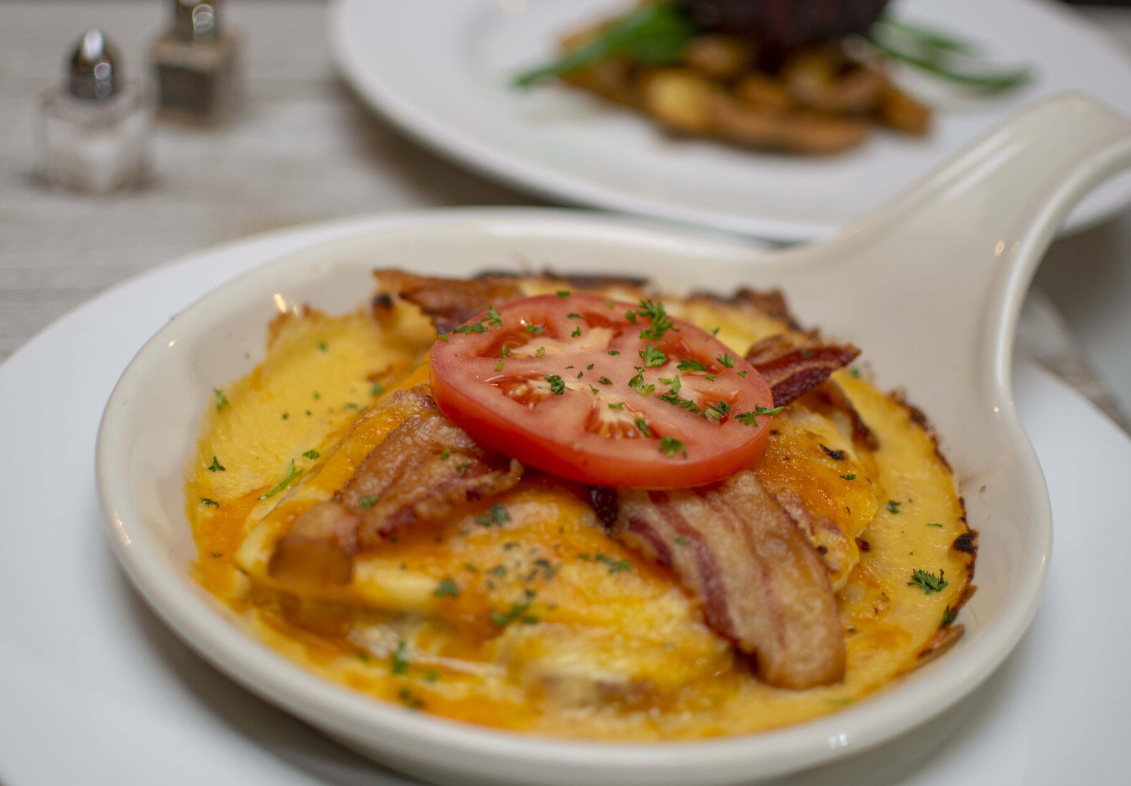 A white ceramic dish containing a breakfast dish with eggs, bacon, and a tomato slice, garnished with herbs.