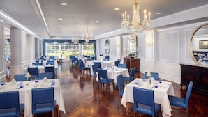 Un comedor formal con mesas dispuestas para el servicio, sillas azules, candelabros y vistas amplias a través de grandes ventanales.
