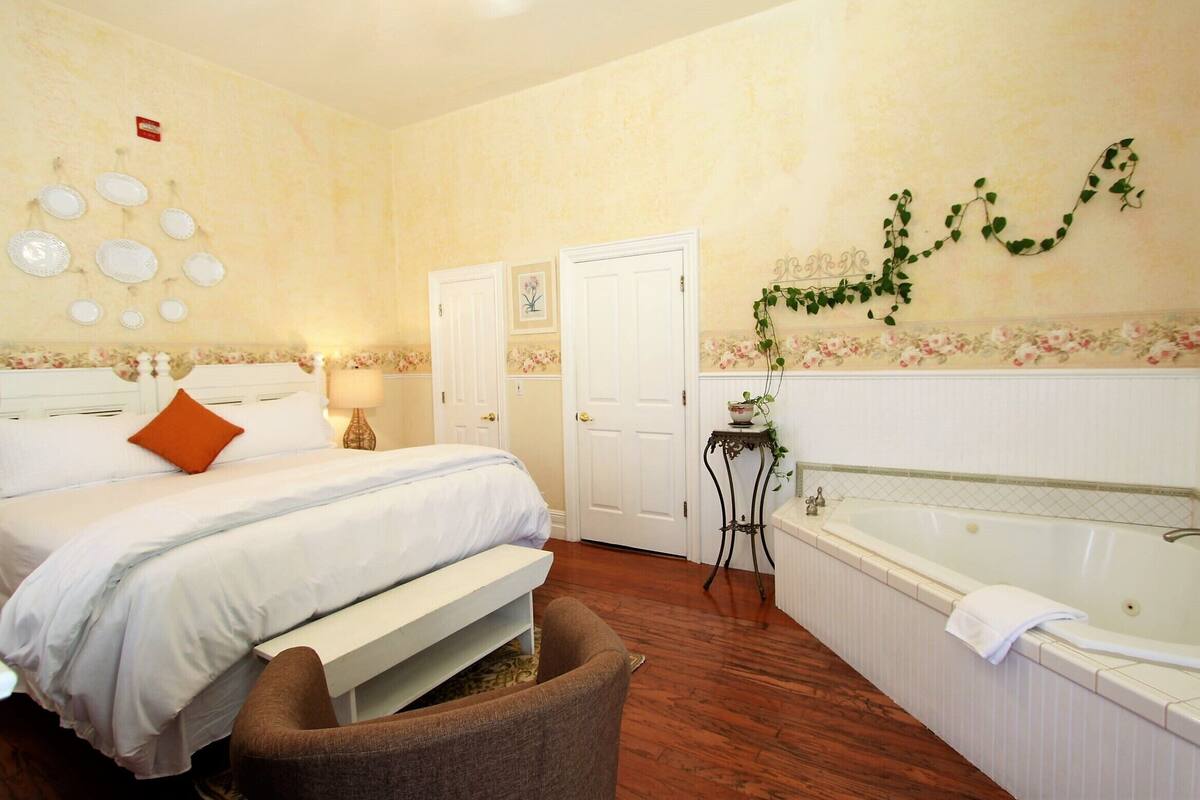 A bedroom with a bed, a bench, a bedside table, a lamp, a door, and a bathroom with a bathtub.
