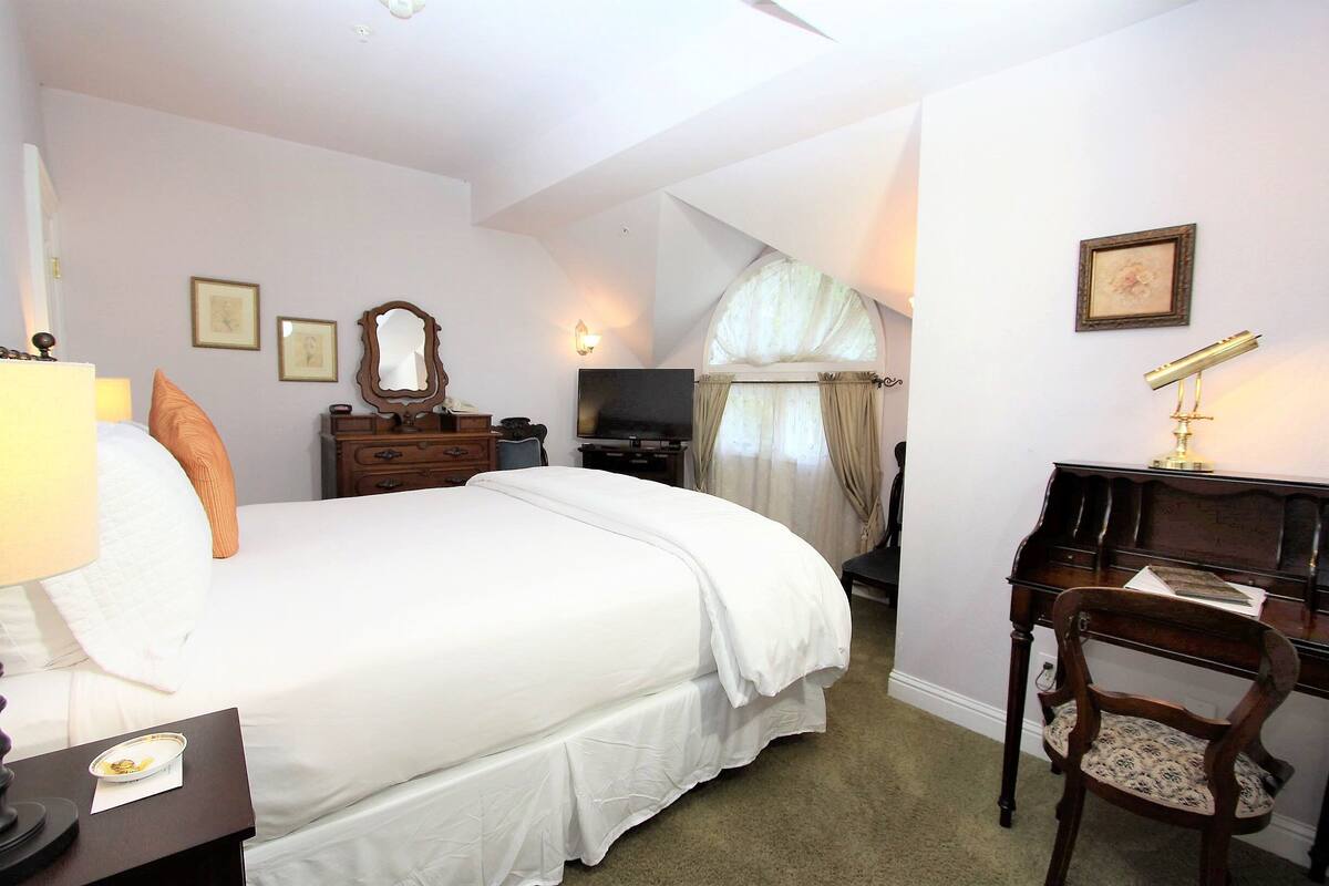 A bedroom with a large bed, a desk with a chair, a television, and a window with curtains.