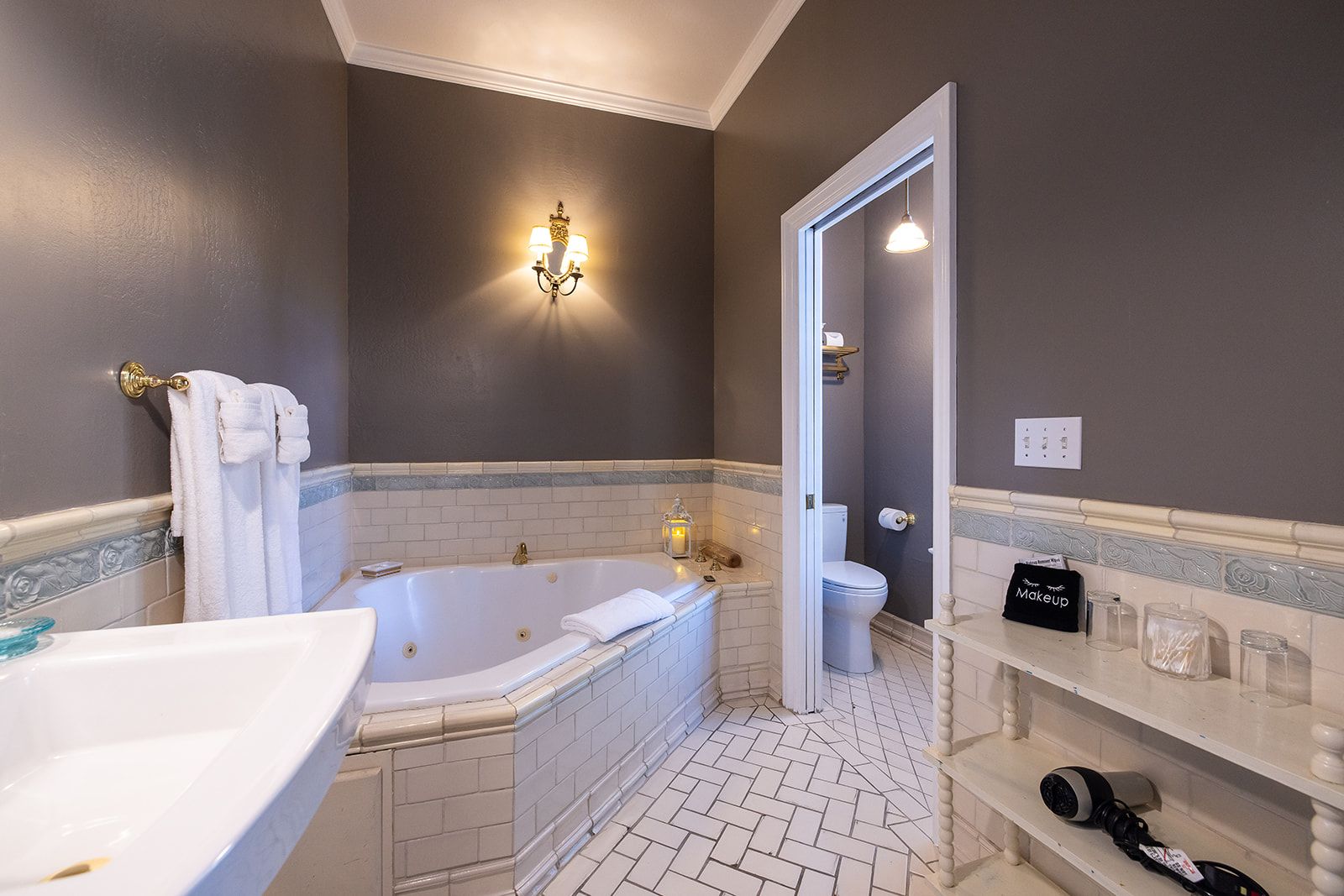 A bathroom with a bathtub, toilet, and sink, featuring a mirror, towel rack, and a shelf with toiletries.