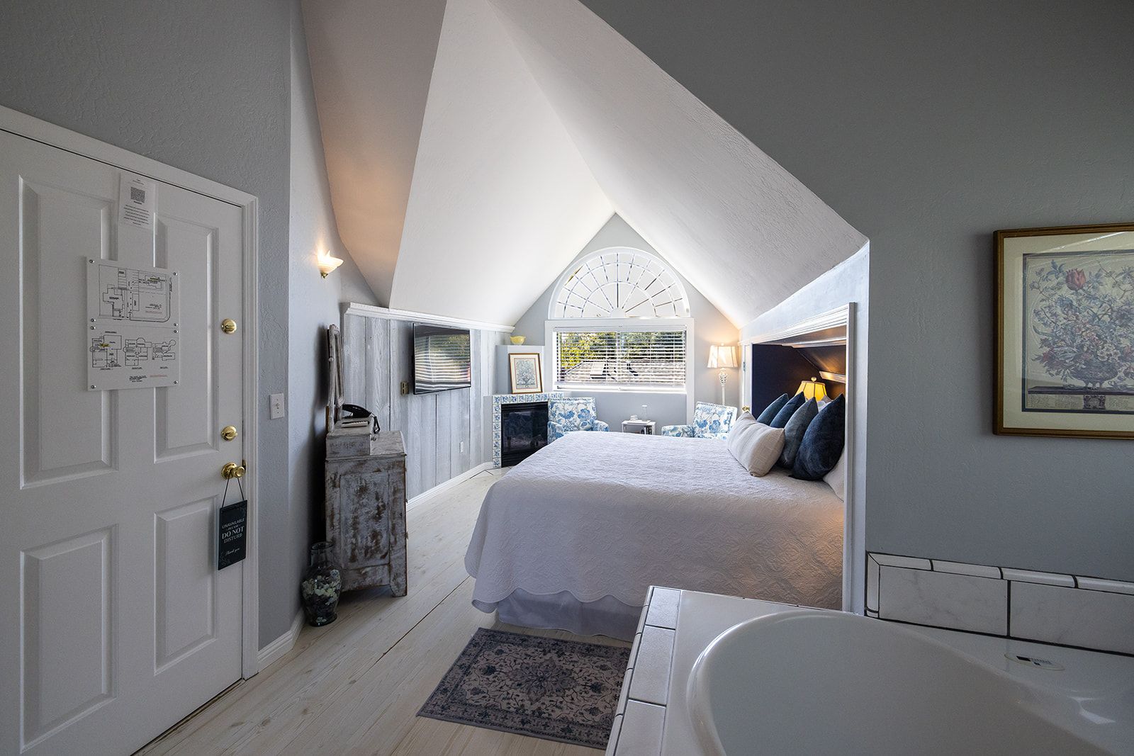 A bedroom with a large bed, a fireplace, a window with blinds, and a bathroom with a bathtub.