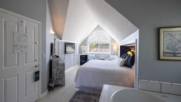 A bedroom with a large bed, a fireplace, a window with blinds, and a bathroom with a bathtub.