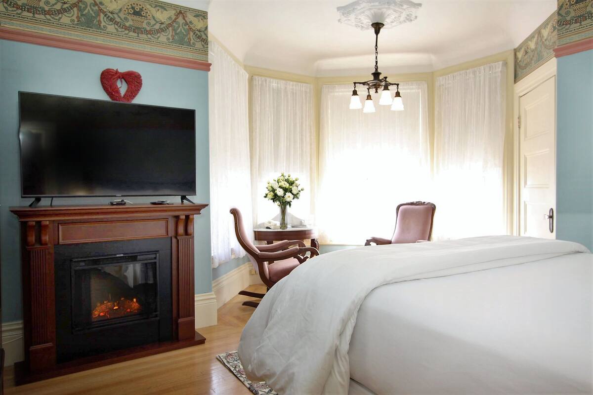 A bedroom with a fireplace, a television, a bed, a chair, and a window with curtains.
