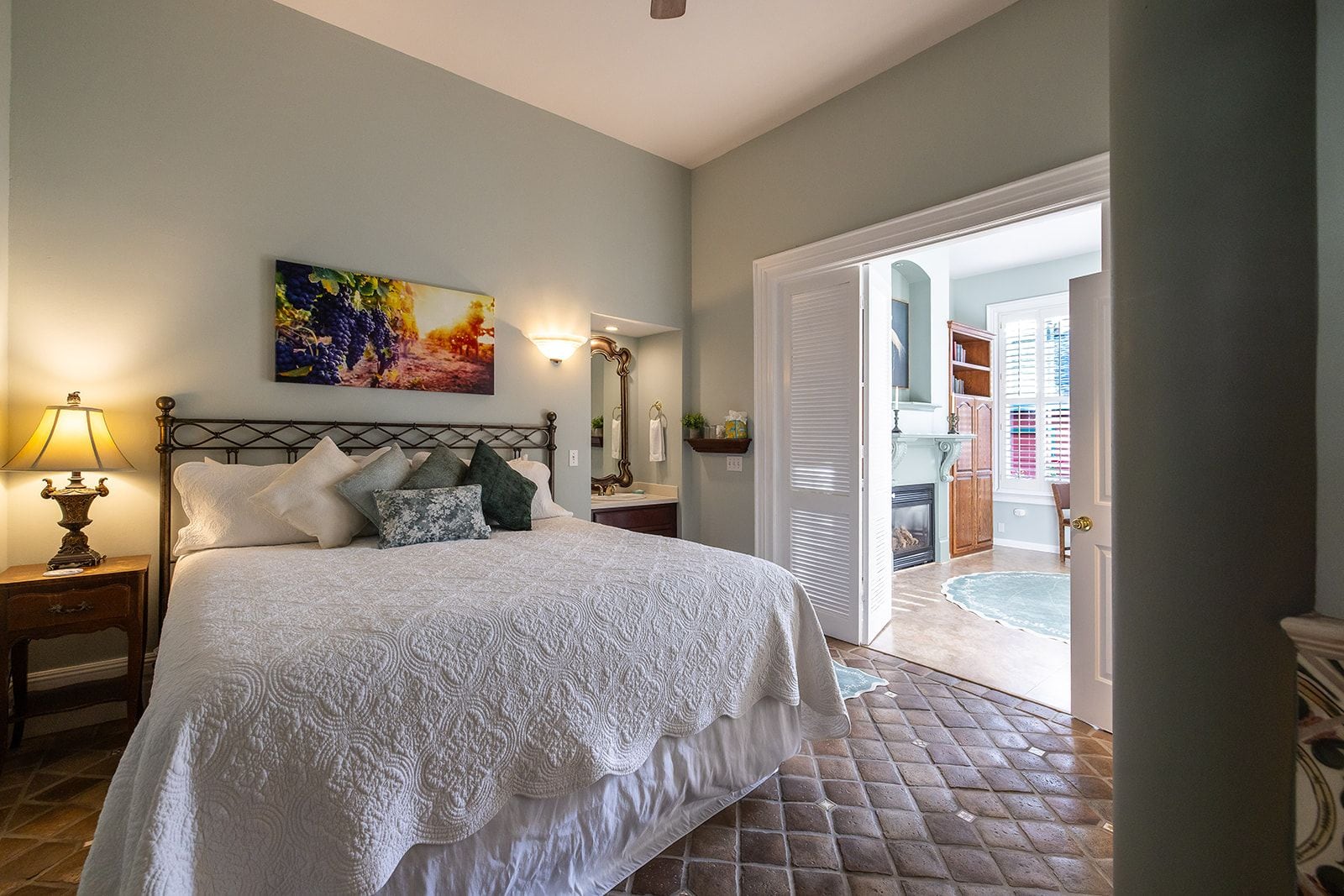 A bedroom with a bed, a nightstand, a lamp, a painting, and a bathroom visible through an open door.