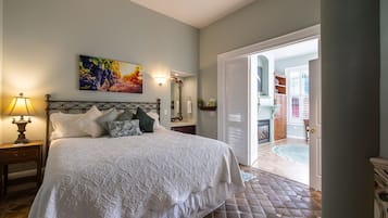 A bedroom with a bed, a nightstand, a lamp, a painting, and a bathroom visible through an open door.