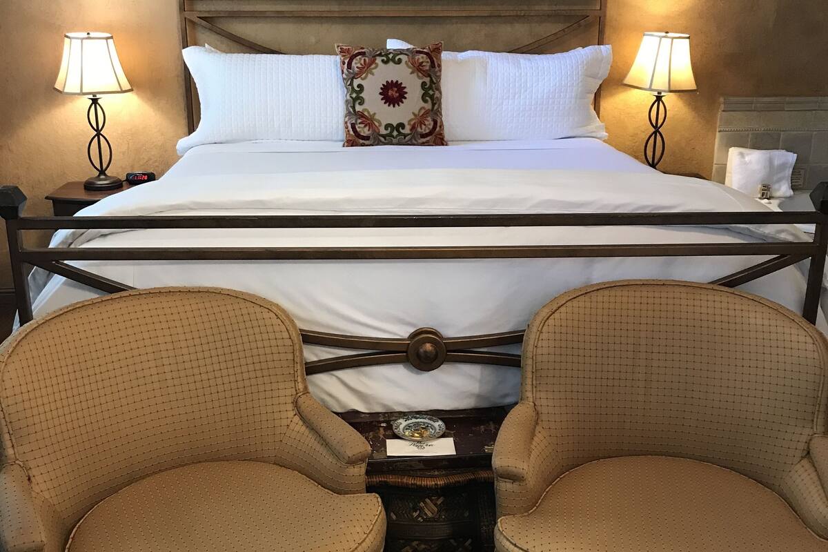 A neatly made bed with white linens, two bedside lamps, and a decorative pillow. There are two wicker chairs in front of the bed.