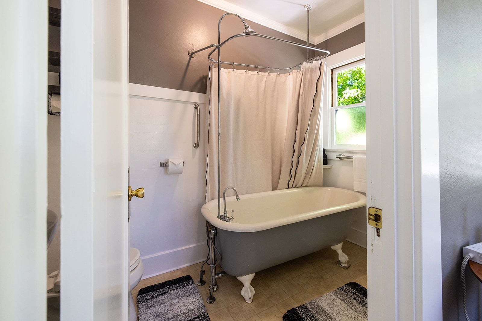 A bathroom with a clawfoot bathtub, a shower curtain, a toilet, and a window with a view of greenery.