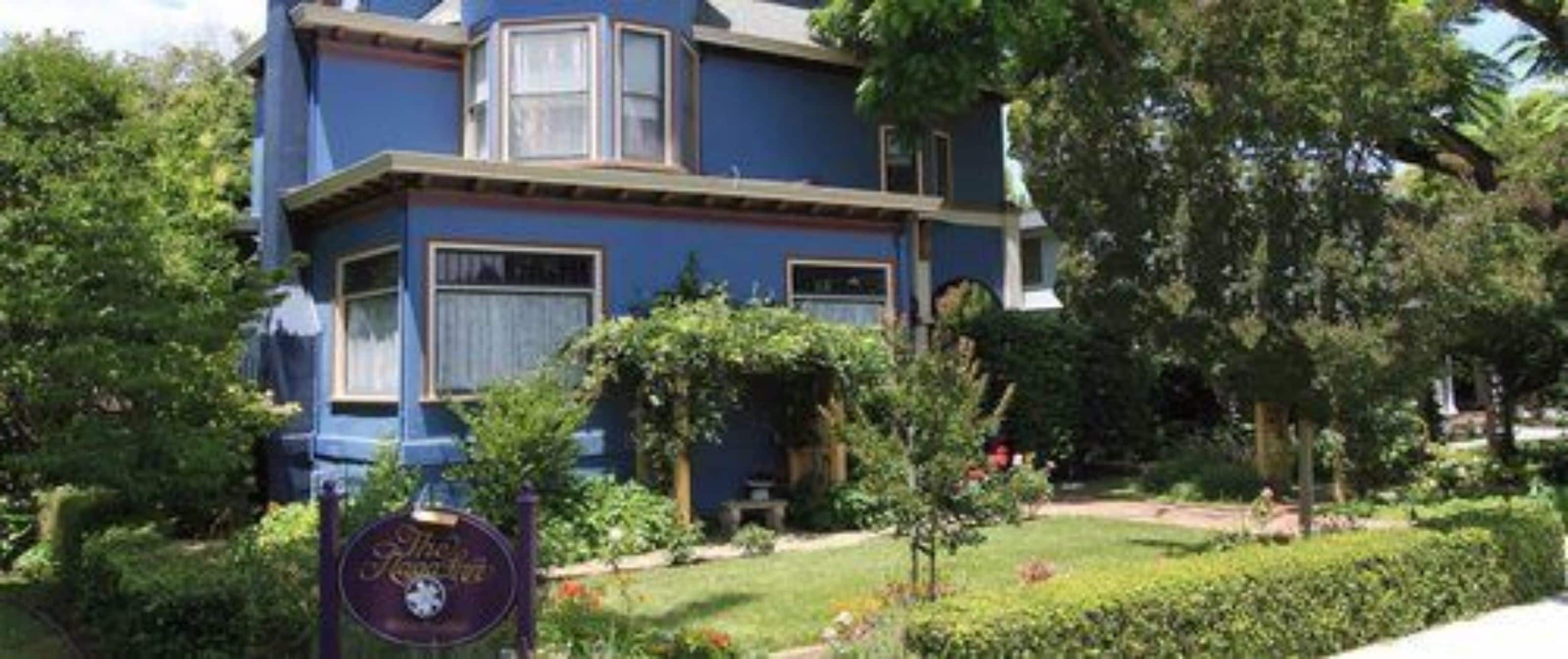 Una casa de dos pisos con fachada azul, rodeada de abundante vegetación.