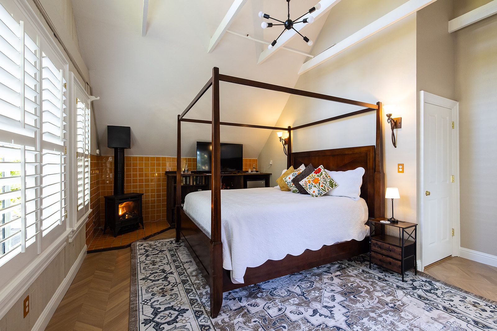 A four-poster bed with a white bedspread and decorative pillows, a television on a wooden stand, a fireplace, and a patterned rug.