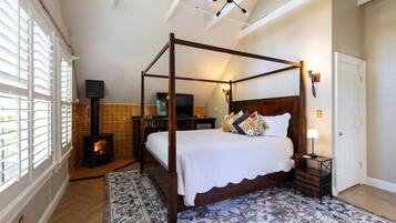 A four-poster bed with a white bedspread and decorative pillows, a television on a wooden stand, a fireplace, and a patterned rug.
