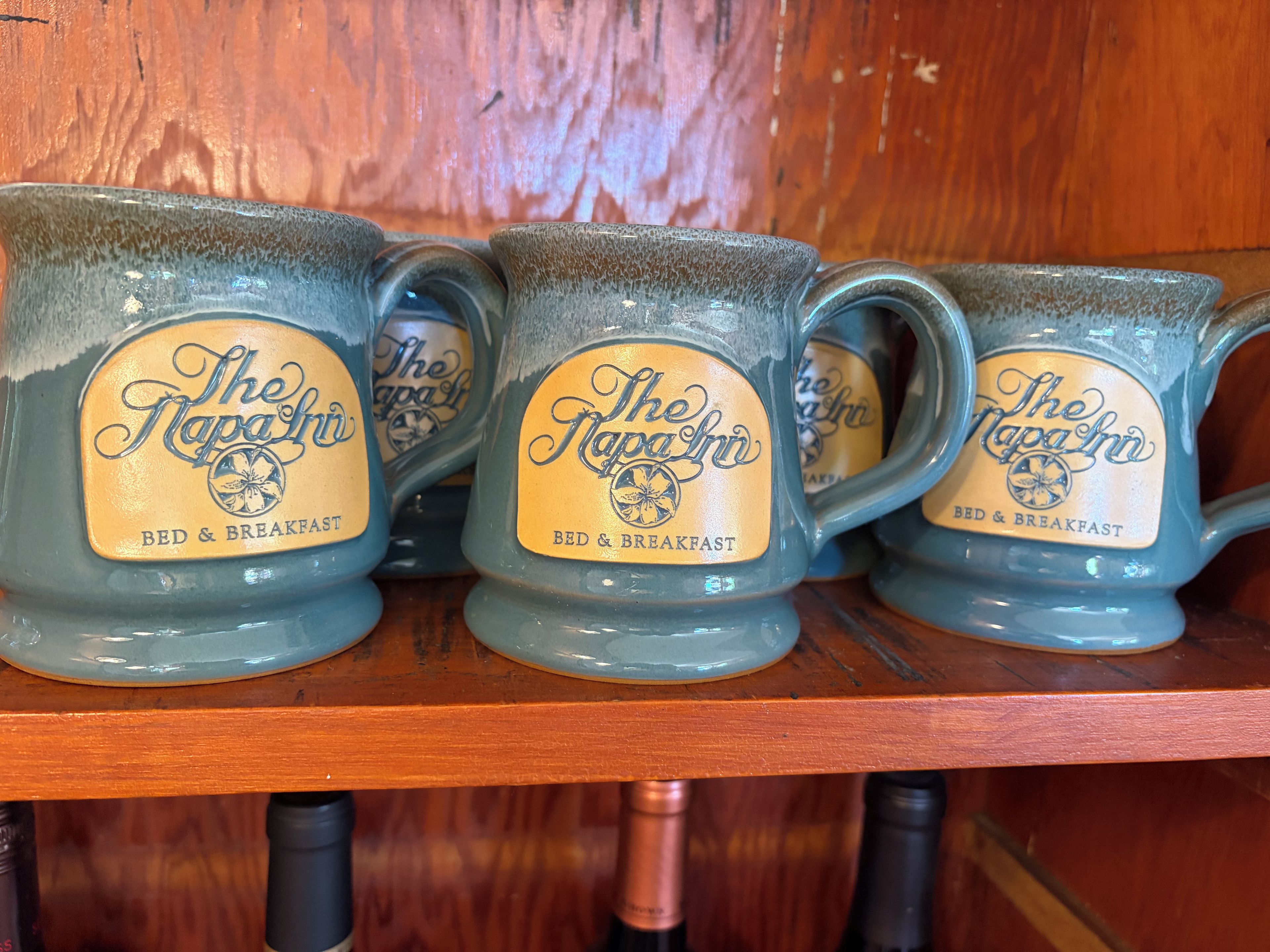 Quatre tasses ornées du logo « The Napa Inn » sont posées sur une étagère en bois.