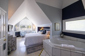Un dormitorio con una cama grande, una zona de estar con una silla, una chimenea, un ventanal con persianas y una bañera.