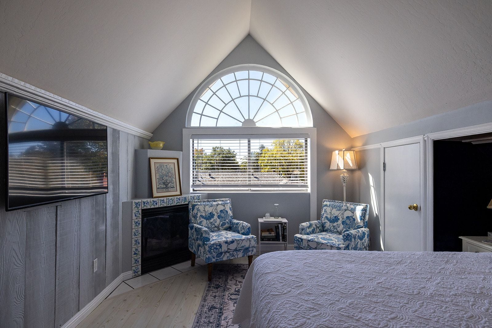 Un dormitorio con chimenea, dos sillones estampados, cama, televisión y ventana con persianas.