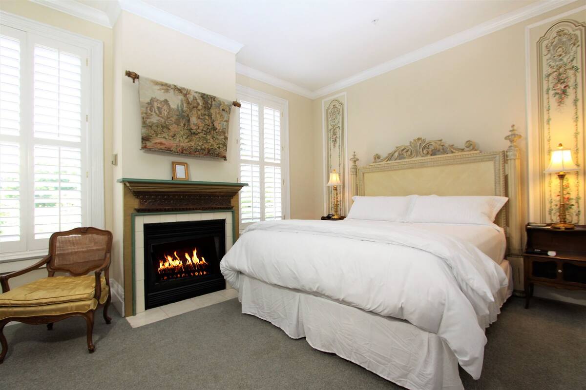 A bedroom with a fireplace, a large bed, a chair, and a nightstand with a lamp.