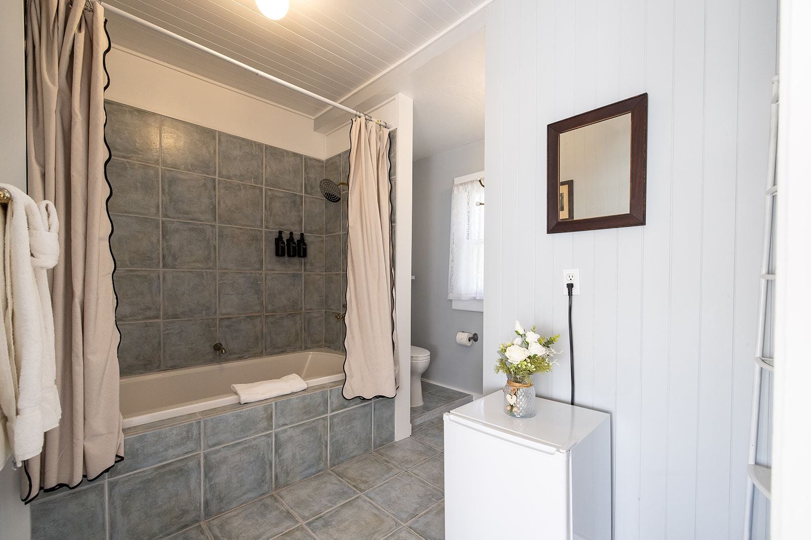 Un baño con bañera, inodoro, lavamanos con un jarrón de flores y un espejo en la pared.