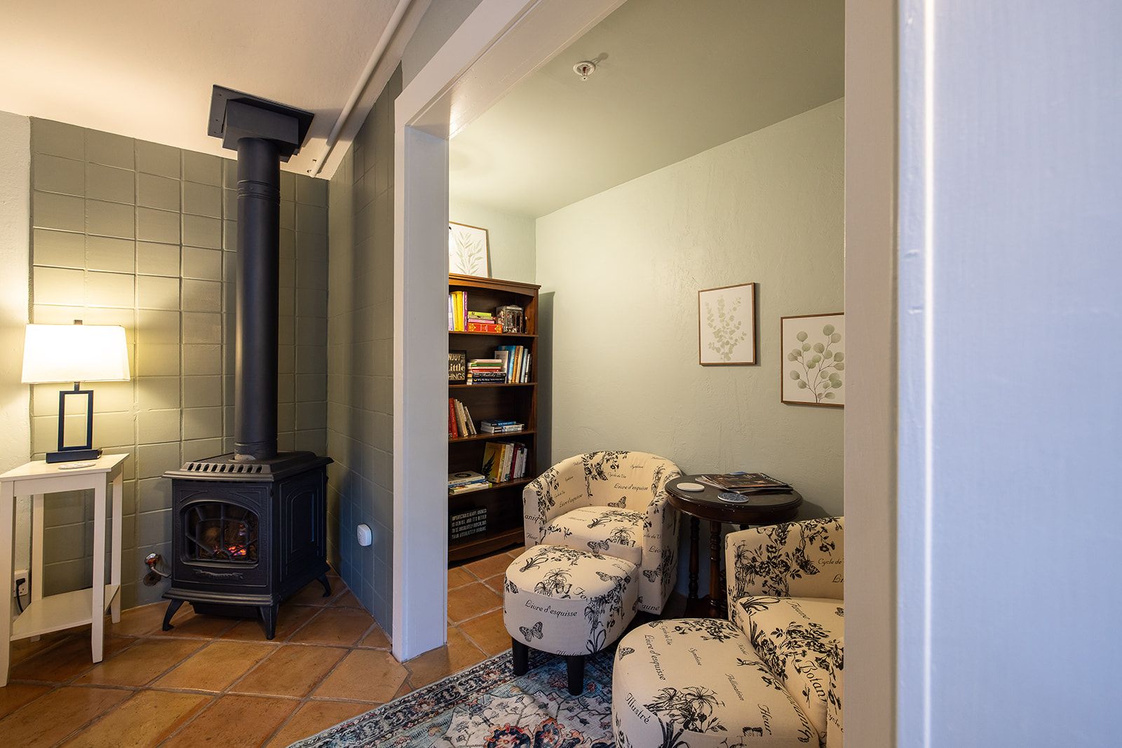 A cozy living room with a fireplace, a bookshelf, floral-patterned furniture, and a patterned rug.