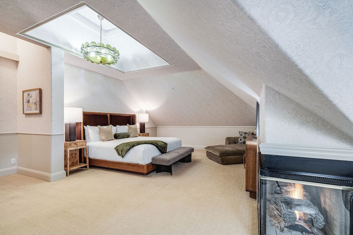 A bedroom with a skylight, a bed, a bench, a fireplace, and a side table.