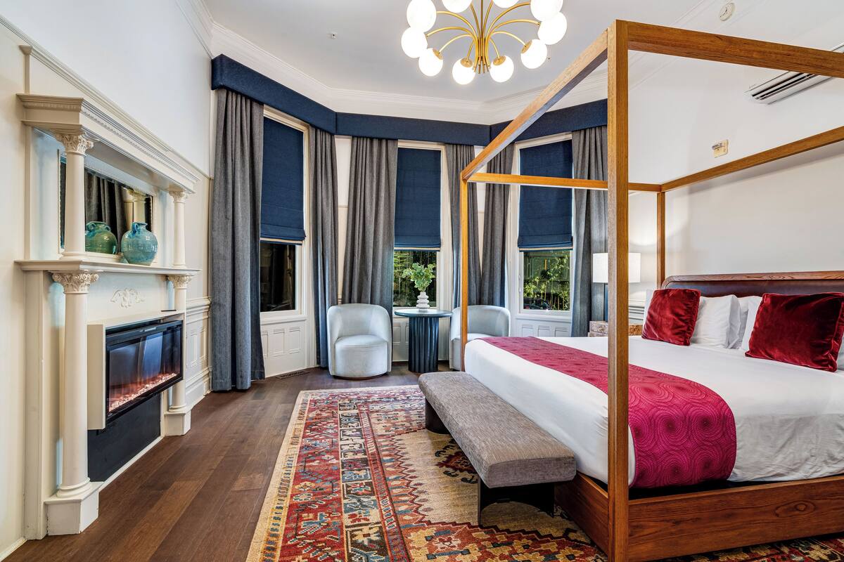 A four-poster bed with a red bedspread, a fireplace with decorative vases, a round table, and a bench.