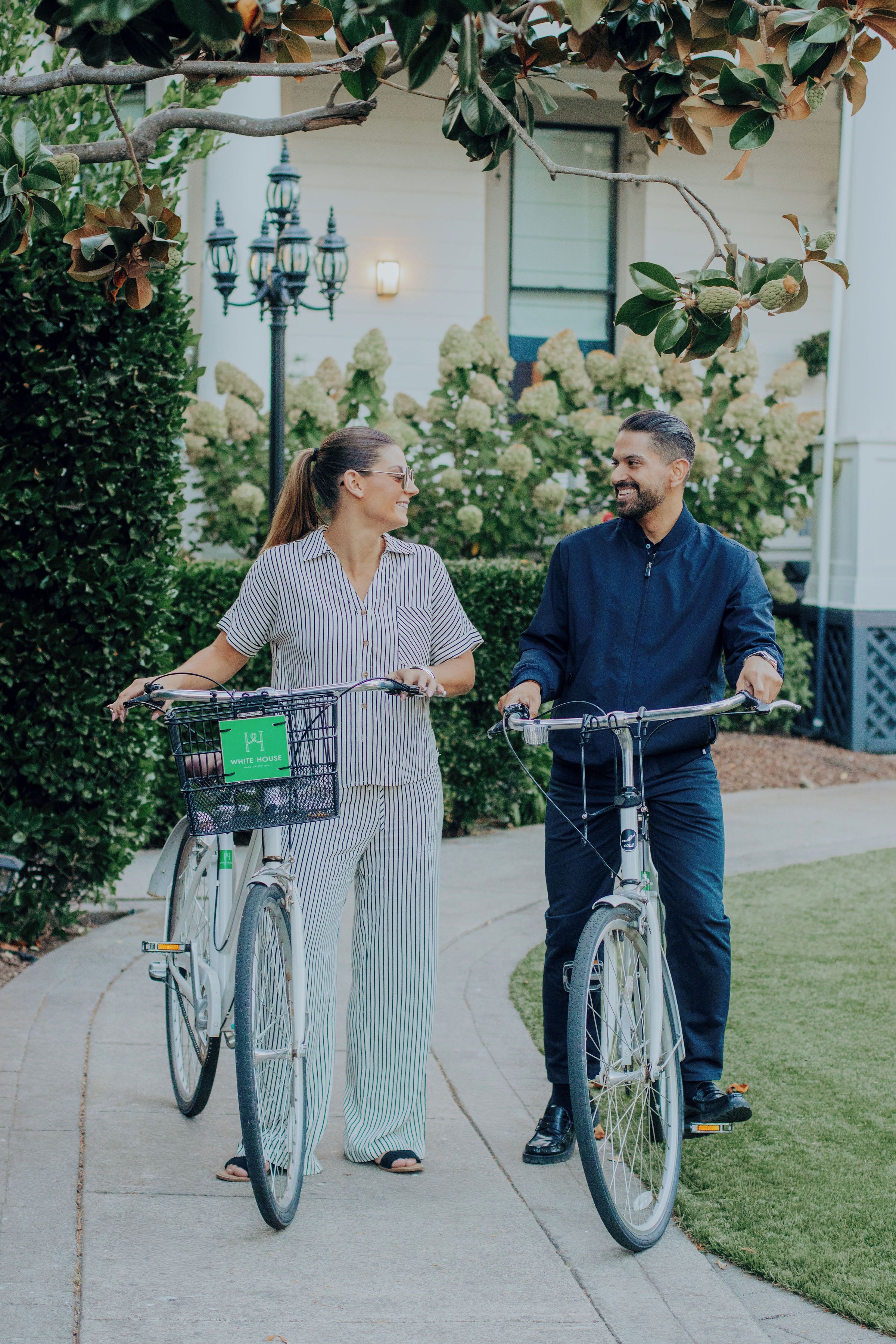 Two people on bicycles with baskets, walking on a sidewalk.