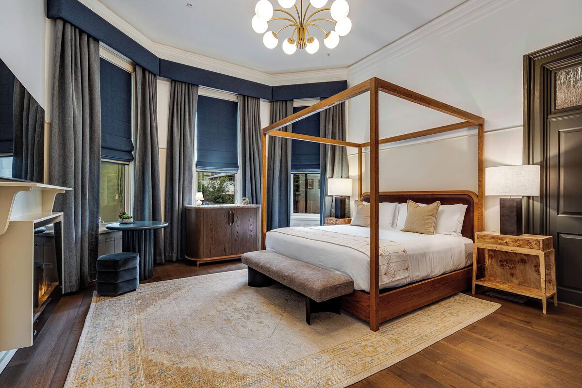 A four-poster bed with a bench, a bedside table, a lamp, and a view of the outdoors through large windows.
