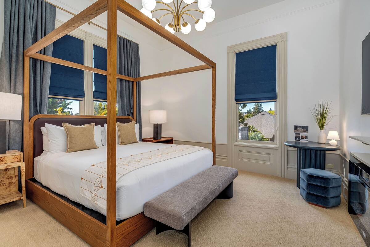A four-poster canopy bed with a bench, bedside tables, a desk with a lamp, and a view of the outside through a window.